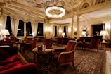 An opulent and elegantly furnished room featuring intricate ceiling designs, grand chandeliers, and plush furnishings. The space is adorned with rich red armchairs and sofas, surrounded by wooden tables. Tall windows are draped with luxurious curtains, providing an ambiance of sophistication. The room is softly lit with table lamps, creating a warm and inviting atmosphere.
