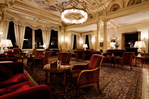 An opulent and elegantly furnished room featuring intricate ceiling designs, grand chandeliers, and plush furnishings. The space is adorned with rich red armchairs and sofas, surrounded by wooden tables. Tall windows are draped with luxurious curtains, providing an ambiance of sophistication. The room is softly lit with table lamps, creating a warm and inviting atmosphere.