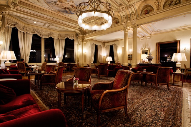 An opulent and elegantly furnished room featuring intricate ceiling designs, grand chandeliers, and plush furnishings. The space is adorned with rich red armchairs and sofas, surrounded by wooden tables. Tall windows are draped with luxurious curtains, providing an ambiance of sophistication. The room is softly lit with table lamps, creating a warm and inviting atmosphere.