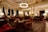 An opulent and elegantly furnished room featuring intricate ceiling designs, grand chandeliers, and plush furnishings. The space is adorned with rich red armchairs and sofas, surrounded by wooden tables. Tall windows are draped with luxurious curtains, providing an ambiance of sophistication. The room is softly lit with table lamps, creating a warm and inviting atmosphere.