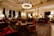 An opulent and elegantly furnished room featuring intricate ceiling designs, grand chandeliers, and plush furnishings. The space is adorned with rich red armchairs and sofas, surrounded by wooden tables. Tall windows are draped with luxurious curtains, providing an ambiance of sophistication. The room is softly lit with table lamps, creating a warm and inviting atmosphere.