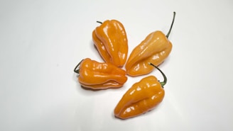 Four bright orange habanero peppers with a glossy surface are arranged on a plain white background. Their uneven, wrinkled textures and green stems suggest freshness.
