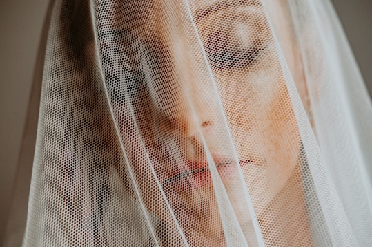 A close-up of a person's face partially covered by a sheer, white veil. The veil adds a textured overlay while soft light highlights the face, creating an ethereal and intimate atmosphere.