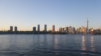 A skyline of a city featuring a prominent tower surrounded by various high-rise buildings. The foreground is dominated by a body of water, reflecting the soft hues of a sunset. The clear sky has a gentle gradient from light blue to warm orange.