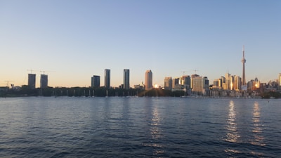 A skyline of a city featuring a prominent tower surrounded by various high-rise buildings. The foreground is dominated by a body of water, reflecting the soft hues of a sunset. The clear sky has a gentle gradient from light blue to warm orange.