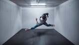 A person is performing an acrobatic jump or dance move mid-air in a small, empty room with concrete walls and a ceiling light. The figure is wearing casual clothing and appears to be in a dynamic pose, creating a sense of motion and fluidity.