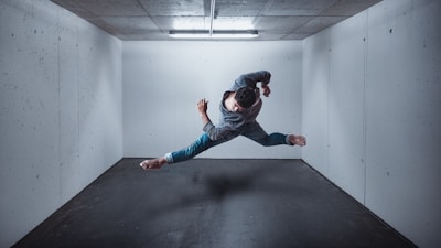 A person is performing an acrobatic jump or dance move mid-air in a small, empty room with concrete walls and a ceiling light. The figure is wearing casual clothing and appears to be in a dynamic pose, creating a sense of motion and fluidity.