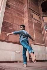 A person is performing a graceful dance move, standing on one leg with the other leg extended behind. The setting features a backdrop of an ancient, ornately designed stone structure, possibly an architectural monument, with large, carved blocks in warm earth tones.