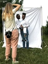 A photographer is capturing a couple posing in front of a white backdrop outdoors. The photographer is wearing casual clothes and has a camera strapped around their neck. The couple, both dressed in white shirts and jeans, stand closely, with the woman resting her hands on her abdomen. The backdrop is set in a grassy area with trees in the background.