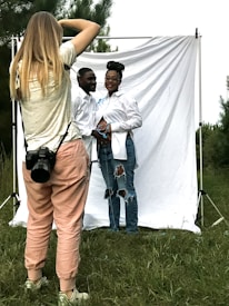 A photographer is capturing a couple posing in front of a white backdrop outdoors. The photographer is wearing casual clothes and has a camera strapped around their neck. The couple, both dressed in white shirts and jeans, stand closely, with the woman resting her hands on her abdomen. The backdrop is set in a grassy area with trees in the background.