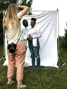 A photographer is capturing a couple posing in front of a white backdrop outdoors. The photographer is wearing casual clothes and has a camera strapped around their neck. The couple, both dressed in white shirts and jeans, stand closely, with the woman resting her hands on her abdomen. The backdrop is set in a grassy area with trees in the background.