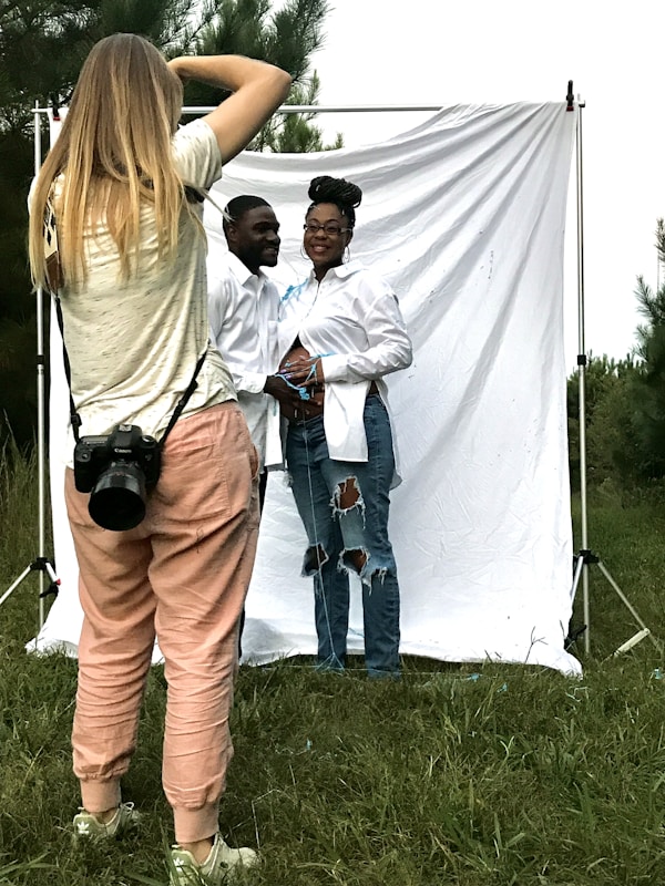 A photographer is capturing a couple posing in front of a white backdrop outdoors. The photographer is wearing casual clothes and has a camera strapped around their neck. The couple, both dressed in white shirts and jeans, stand closely, with the woman resting her hands on her abdomen. The backdrop is set in a grassy area with trees in the background.