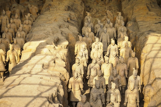 A vast assembly of ancient terracotta warriors, standing in rows within an excavated pit. The figures are intricately detailed, depicting soldiers in battle attire, crafted from clay and showing signs of weathering. The lighting casts soft shadows across the scene, highlighting the stone-like texture of the statues.