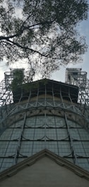 A steel structure with large, grid-like windows reaches up towards the sky, flanked by two tower-like elements. Overhead, tree branches with leaves partially obscure the view, adding a natural contrast to the industrial feel of the metal framework.