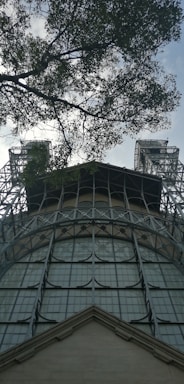 A steel structure with large, grid-like windows reaches up towards the sky, flanked by two tower-like elements. Overhead, tree branches with leaves partially obscure the view, adding a natural contrast to the industrial feel of the metal framework.
