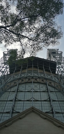A steel structure with large, grid-like windows reaches up towards the sky, flanked by two tower-like elements. Overhead, tree branches with leaves partially obscure the view, adding a natural contrast to the industrial feel of the metal framework.