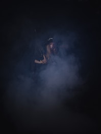 A shadowy figure draped in dark, occult-inspired clothing under dim, moody lighting.