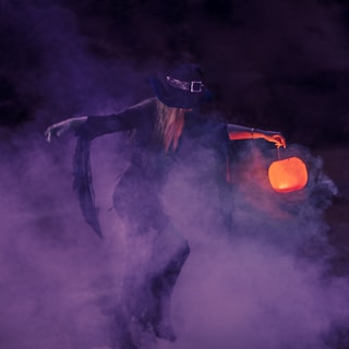 A figure dressed in dark, flowing garments and a wide-brimmed hat, resembling a witch, is surrounded by dense purple fog. In one hand, a glowing orange lantern adds a mysterious light to the scene.