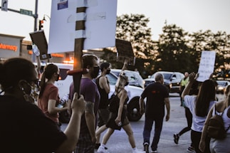 A group of people is participating in a protest, holding up signs in a street. Most participants are wearing face masks. There is a police vehicle with flashing lights visible in the background along with some parked cars and a pharmacy sign.