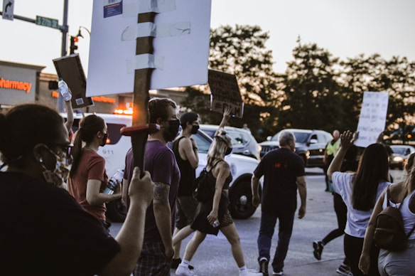 A group of people is participating in a protest, holding up signs in a street. Most participants are wearing face masks. There is a police vehicle with flashing lights visible in the background along with some parked cars and a pharmacy sign.