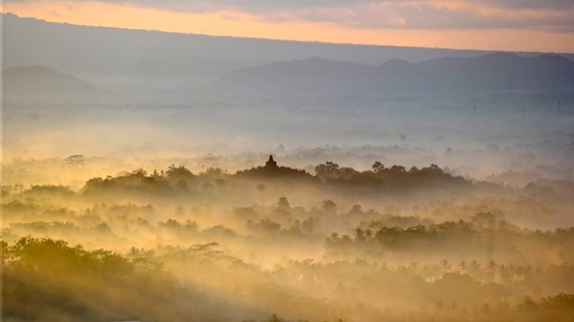 A serene landscape with mist covering lush greenery. In the distance, a silhouette of a temple rises above the trees, surrounded by soft, golden morning light. The sky displays gentle hues of orange and blue as the sun breaks through the clouds.