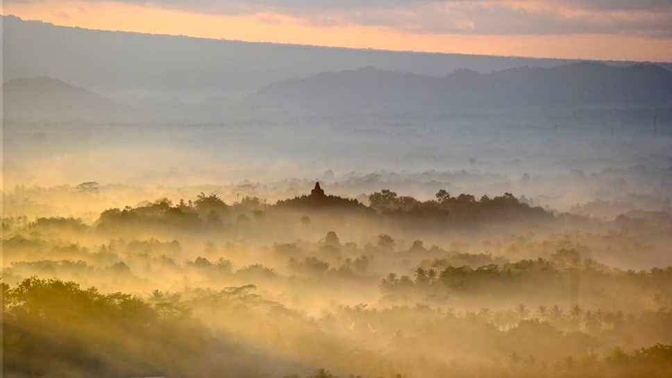 A serene landscape with mist covering lush greenery. In the distance, a silhouette of a temple rises above the trees, surrounded by soft, golden morning light. The sky displays gentle hues of orange and blue as the sun breaks through the clouds.