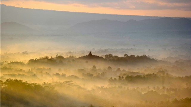 A serene landscape with mist covering lush greenery. In the distance, a silhouette of a temple rises above the trees, surrounded by soft, golden morning light. The sky displays gentle hues of orange and blue as the sun breaks through the clouds.