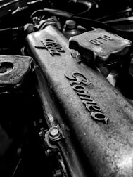 A close-up view of a metal engine component with embossed lettering, depicting a vintage or classic car part with visible details of bolts and textured metal surfaces.
