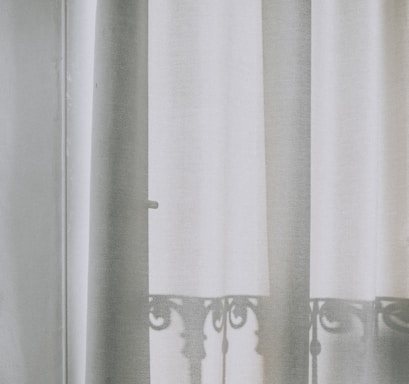 A soft, semi-transparent curtain hangs in front of a window, allowing the light to filter through gently. The outline of an ornate iron balcony railing is visible as a shadow on the curtain, adding a delicate and intricate pattern to the otherwise plain fabric.