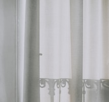 A soft, semi-transparent curtain hangs in front of a window, allowing the light to filter through gently. The outline of an ornate iron balcony railing is visible as a shadow on the curtain, adding a delicate and intricate pattern to the otherwise plain fabric.