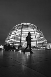 grayscale photo of man in jacket and pants standing in front of glass building
