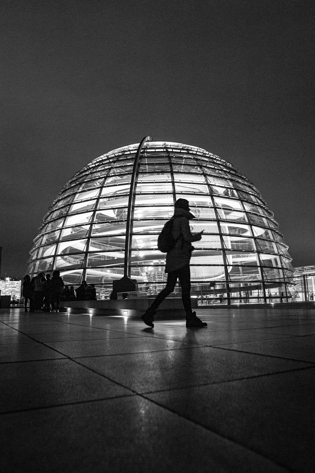 grayscale photo of man in jacket and pants standing in front of glass building