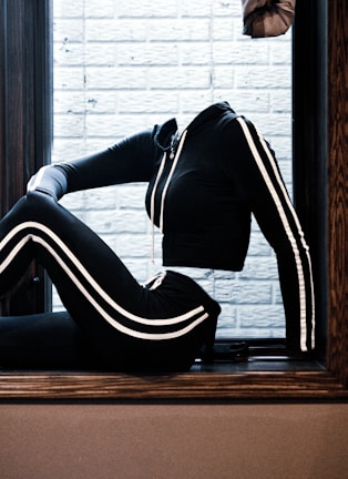 A black hooded tracksuit with white stripes is posed sitting on a window ledge. The background consists of a white brick wall, and no head or feet are visible.