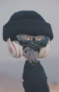 A clever arrangement where the head of a guitar is accessorized with black sunglasses, white headphones, a navy knitted beanie, and a knitted scarf, creating the illusion of a person.