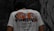 A person is wearing a white t-shirt with a colorful dragon design and the word 'FORTUNE' printed on the back. The shirt also includes text in a foreign language along with motivational phrases such as 'Fate is influenced by strokes of Good Luck as well as Humup Setbacks.' The setting appears to be outdoors with a stone or graffiti-covered wall in the background.