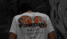 A person is wearing a white t-shirt with a colorful dragon design and the word 'FORTUNE' printed on the back. The shirt also includes text in a foreign language along with motivational phrases such as 'Fate is influenced by strokes of Good Luck as well as Humup Setbacks.' The setting appears to be outdoors with a stone or graffiti-covered wall in the background.
