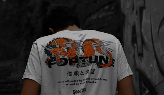 A person is wearing a white t-shirt with a colorful dragon design and the word 'FORTUNE' printed on the back. The shirt also includes text in a foreign language along with motivational phrases such as 'Fate is influenced by strokes of Good Luck as well as Humup Setbacks.' The setting appears to be outdoors with a stone or graffiti-covered wall in the background.