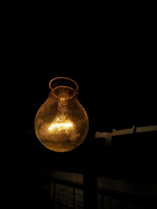 A glowing incandescent light bulb hangs against a dark background. The filament inside the bulb is clearly visible, emitting a warm yellow light. The contrast between the bright bulb and the darkness creates a dramatic effect.
