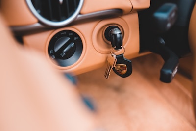 A car interior with tan upholstery featuring a close-up of the ignition area. A black car key with a logo and a few other keys on a ring are inserted in the ignition. The dashboard includes controls and vents.