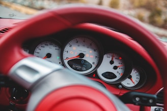 A close-up view of a car dashboard featuring several gauges including speedometer, tachometer, and fuel indicators, all with white backgrounds and black numerals. The steering wheel and surrounding interior are covered in red leather.