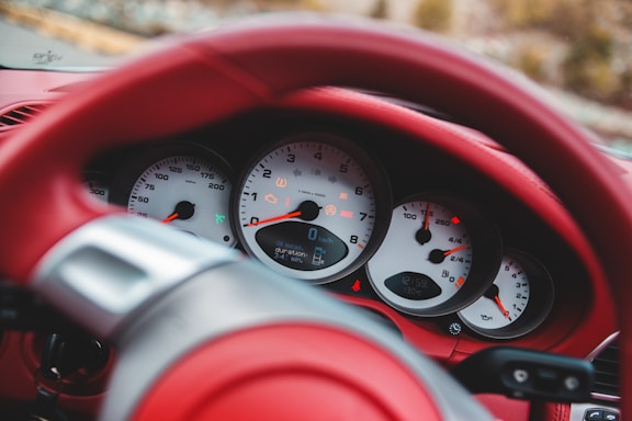 A close-up view of a car dashboard featuring several gauges including speedometer, tachometer, and fuel indicators, all with white backgrounds and black numerals. The steering wheel and surrounding interior are covered in red leather.