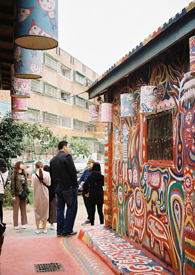 A colorful alley featuring vibrant murals with abstract and animal-like designs covering the walls. A group of people is gathered near the building, observing the artwork. Hanging lanterns with cartoon characters add to the whimsical atmosphere. The background includes a modern building and some greenery.