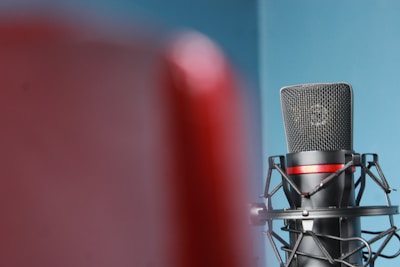 A professional microphone is positioned against a blue background, partially obscured by a red object in the foreground. The microphone is secured in a shock mount, featuring a black and red color scheme.