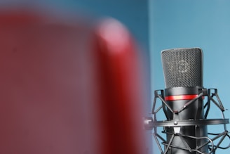A professional microphone is positioned against a blue background, partially obscured by a red object in the foreground. The microphone is secured in a shock mount, featuring a black and red color scheme.