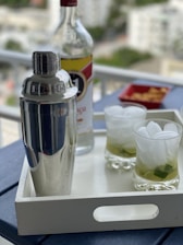 A silver cocktail shaker and a partially full bottle of liquor are placed on a white tray. Two glasses containing a drink with ice and lime are also on the tray, set against a blurred background of a cityscape. Next to the bottle is a small red dish with some snacks.