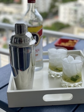 A silver cocktail shaker and a partially full bottle of liquor are placed on a white tray. Two glasses containing a drink with ice and lime are also on the tray, set against a blurred background of a cityscape. Next to the bottle is a small red dish with some snacks.