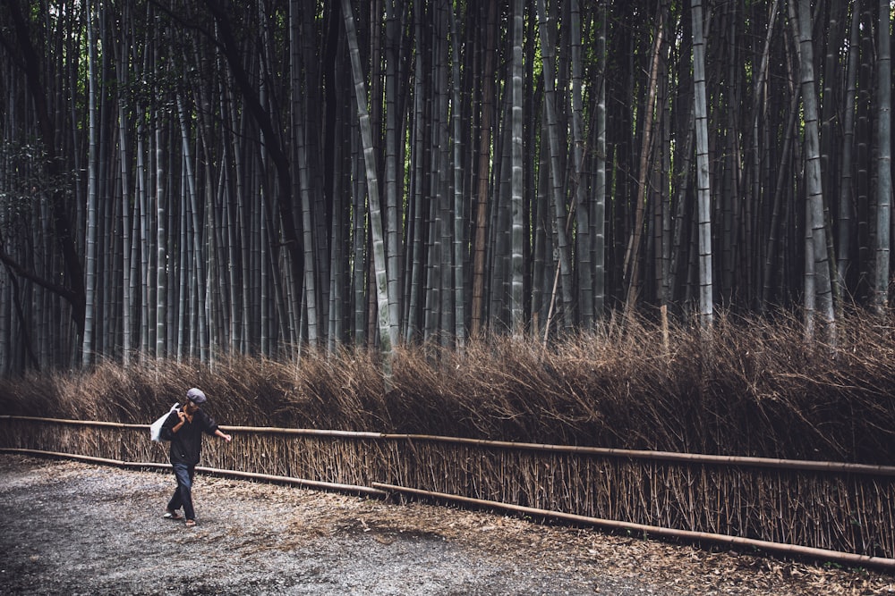 man in black jacket and black pants walking on brown dirt road between green trees during