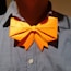 A close-up of a person wearing a bright orange origami bow tie on a grey shirt. The bow tie is prominently folded with angular edges and casts a slight shadow on the shirt.