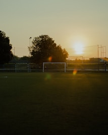 A soccer field is depicted during sunset, with the fading sun casting a warm golden glow over the scene. The goalpost and net are visible in the foreground, with trees and fencing surrounding the area. The sky is clear, enhancing the serene atmosphere.