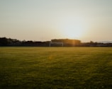 A tranquil football field at sunset, with a goalpost in the distance and lush green grass covering the ground. The sun is low on the horizon, casting a golden glow over the entire scene.